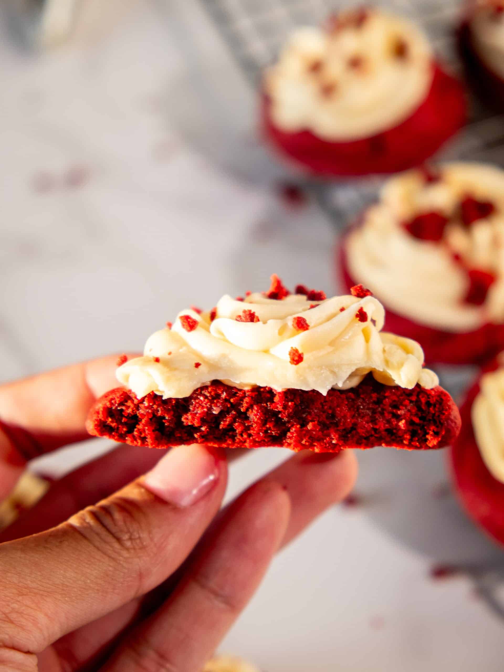 Hand holding a soft red velvet cookie with cream cheese frosting, showing its moist and chewy texture.