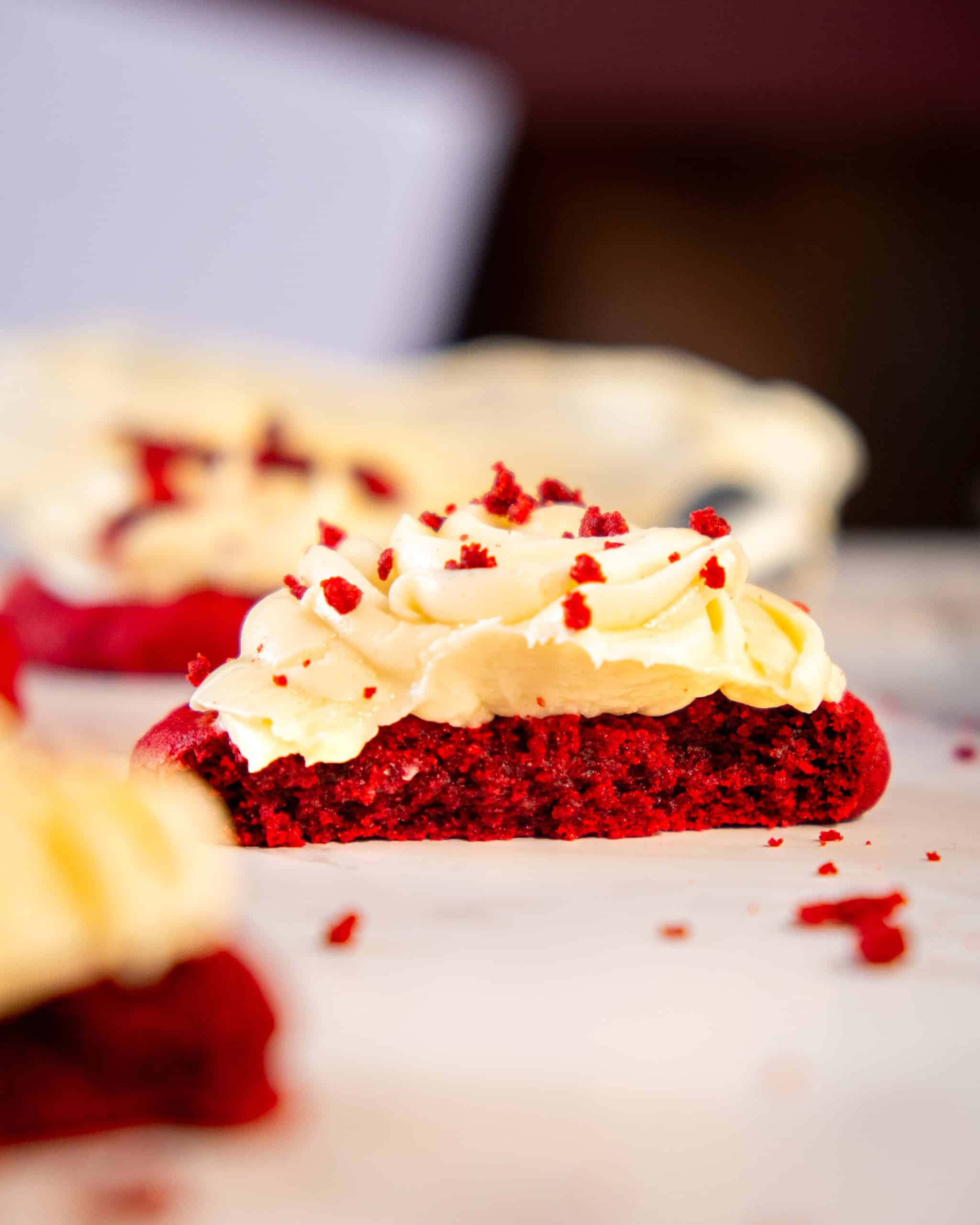 A freshly baked red velvet cookie topped with cream cheese frosting, garnished with red velvet crumbs, highlighting its soft and moist texture.