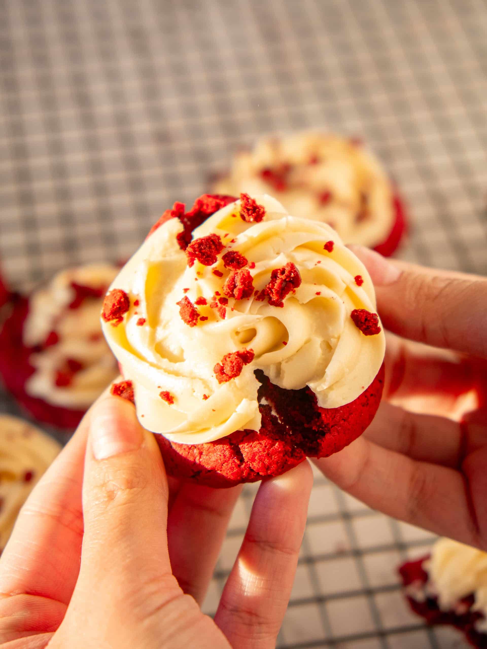 A freshly baked red velvet cookie topped with cream cheese frosting, garnished with red velvet crumbs, highlighting its soft and moist texture.