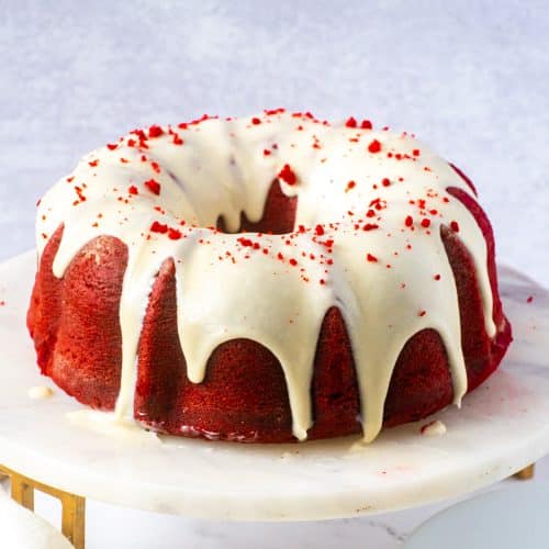 Whole red velvet pound cake with cream cheese frosting on a marble cake stand, topped with red velvet crumbs.