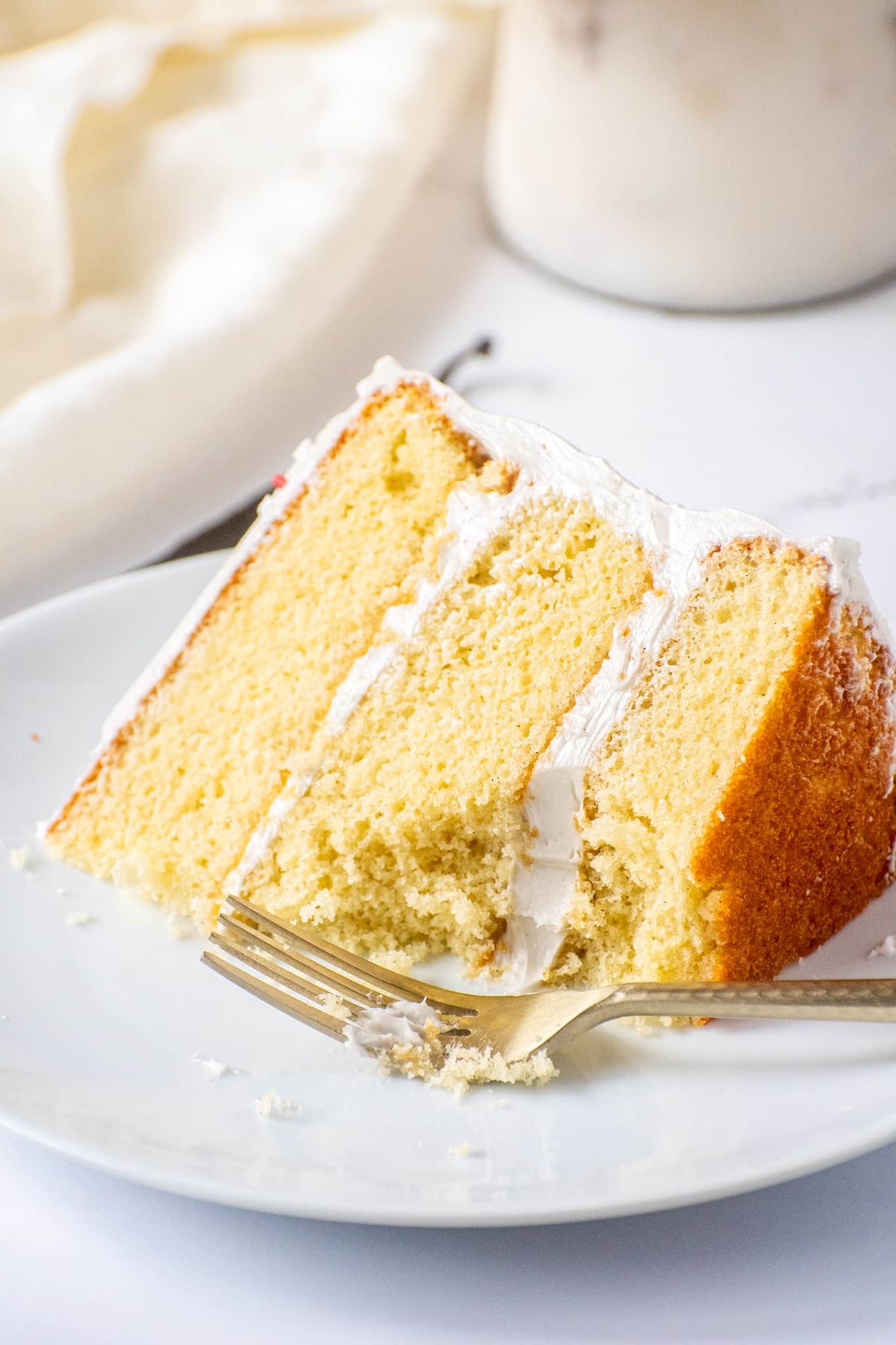 Partially eaten slice of moist vanilla cake with fluffy frosting on a white plate and gold fork.