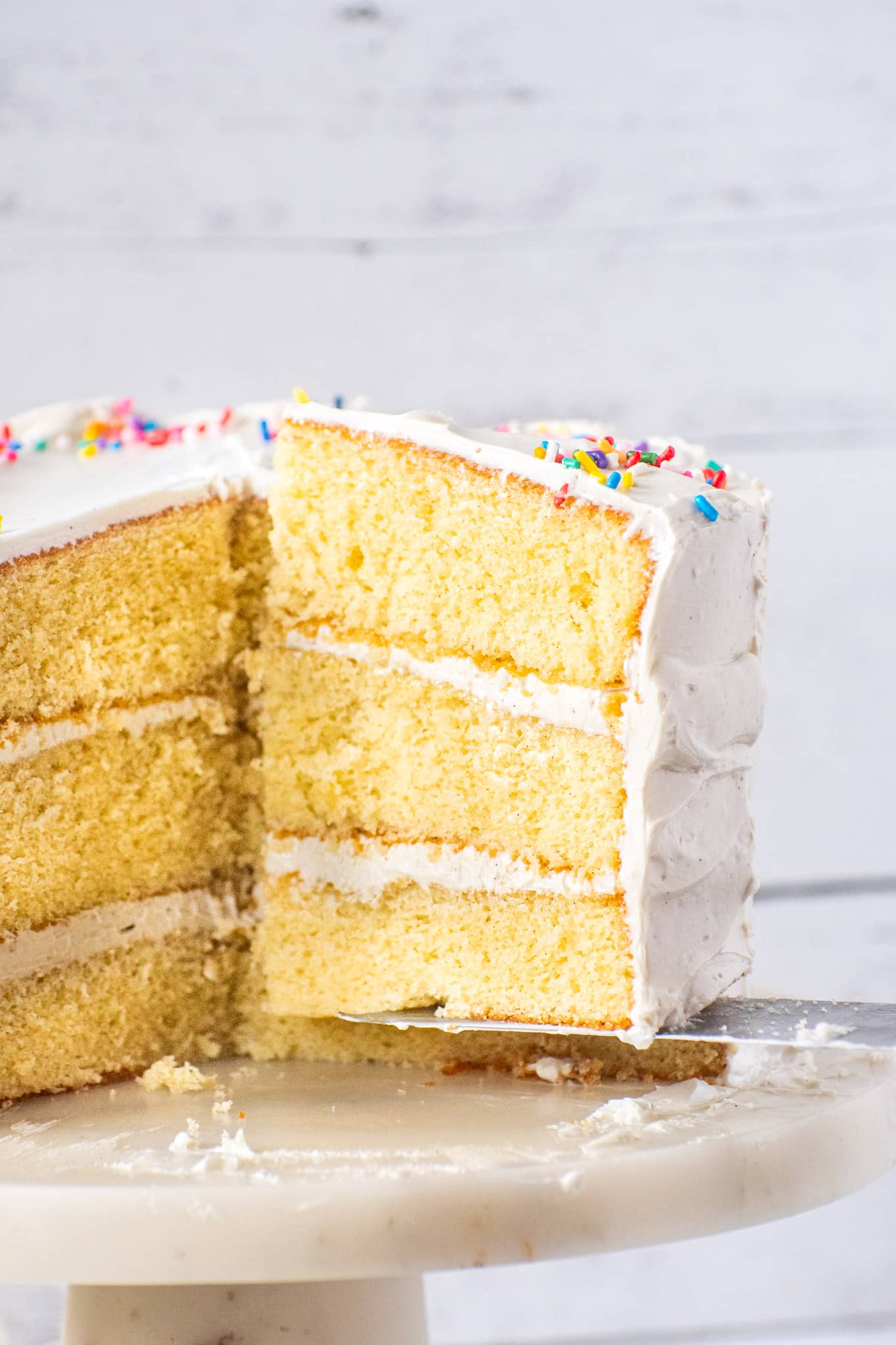 Three-layer vanilla cake with white buttercream frosting and rainbow sprinkles, with a slice removed to show the soft interior.