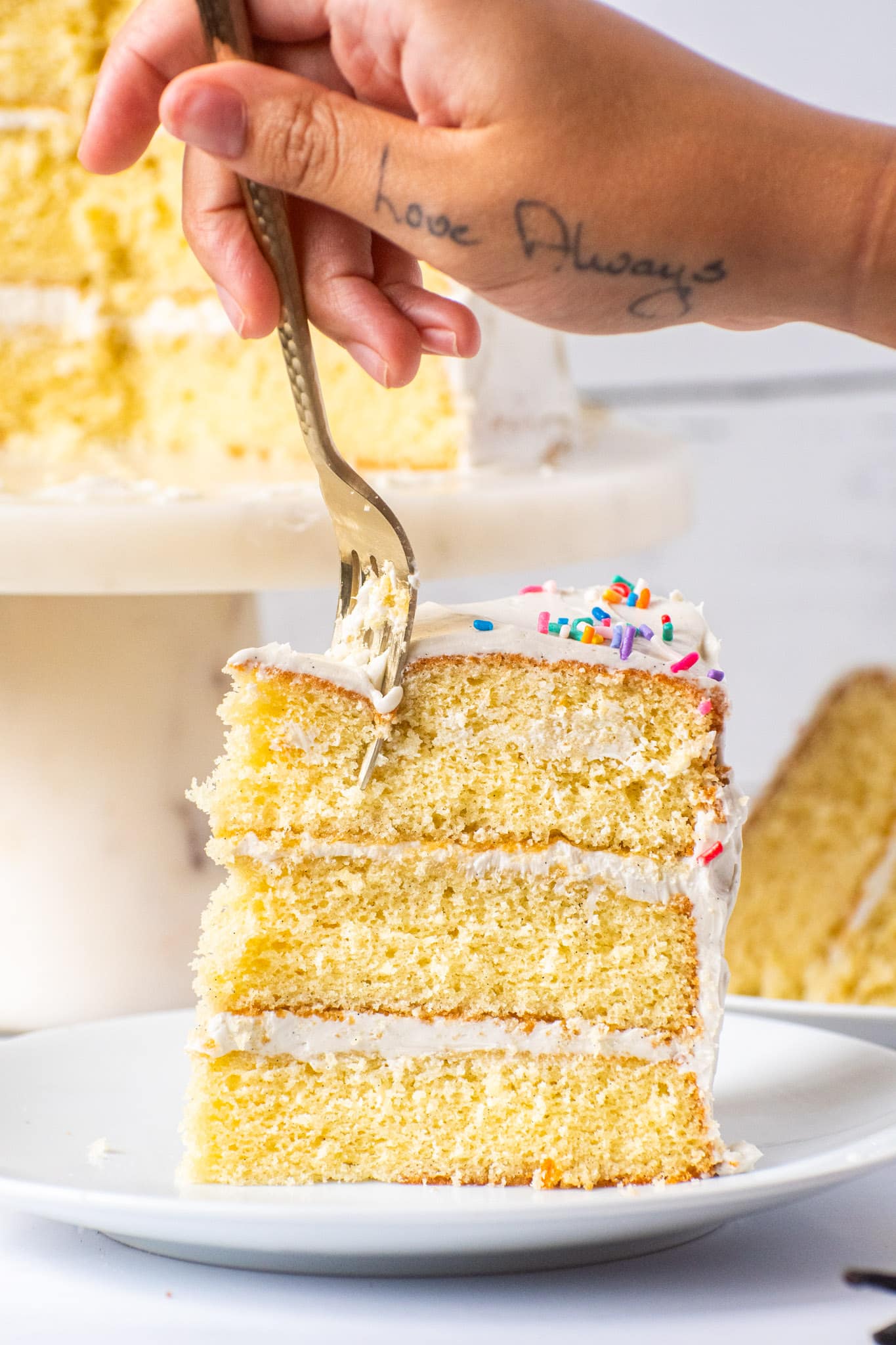 Slice of moist vanilla cake with white frosting and rainbow sprinkles, with a fork taking a bite.
