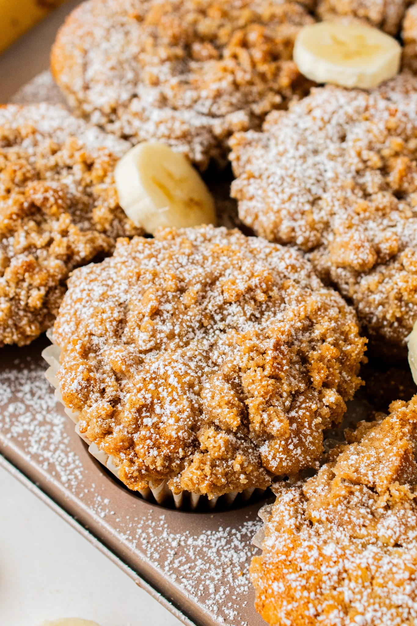 Close-up of a golden banana crumb muffin topped with buttery cinnamon crumble and powdered sugar, showing the crisp, bakery-style texture.