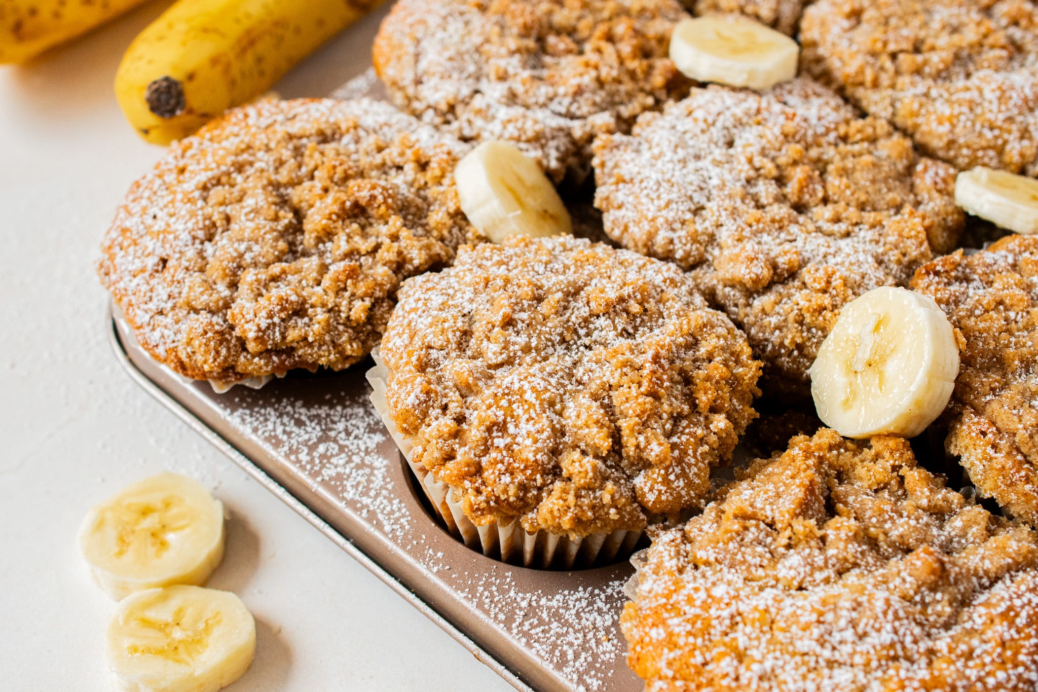 Freshly baked banana crumb muffins topped with buttery cinnamon crumble and dusted with powdered sugar, arranged on a baking pan with banana slices.