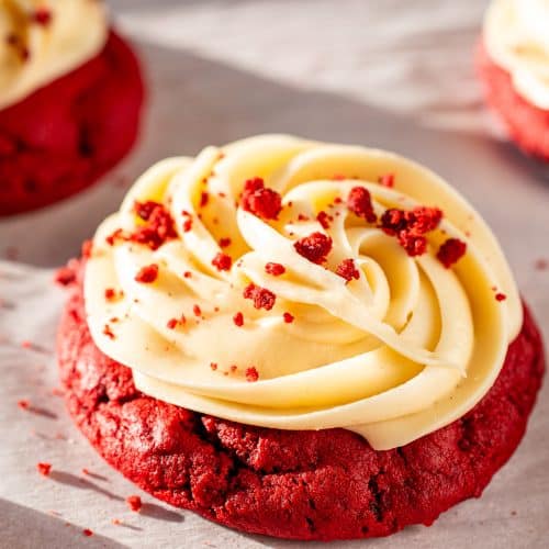 Close-up of a red velvet cookie with cream cheese frosting, topped with red velvet crumbs on parchment paper.