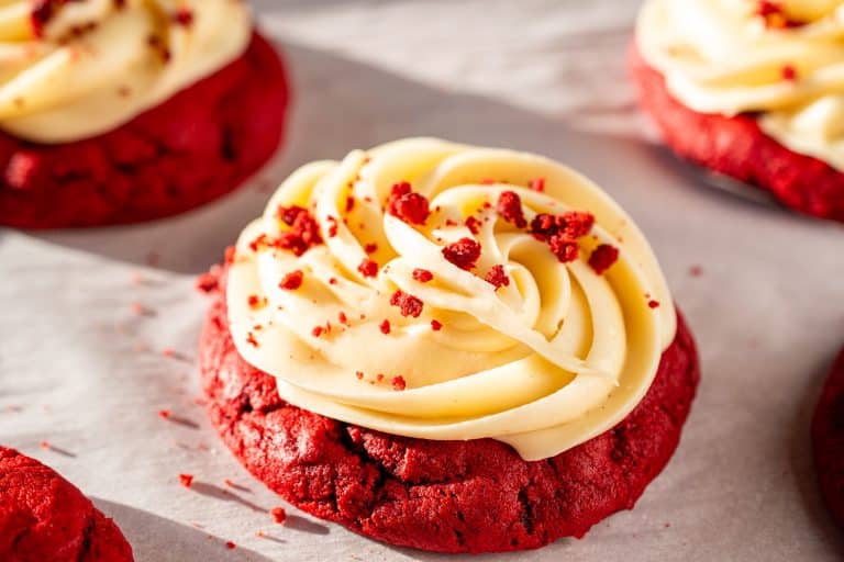 Close-up of a red velvet cookie with cream cheese frosting, topped with red velvet crumbs on parchment paper.