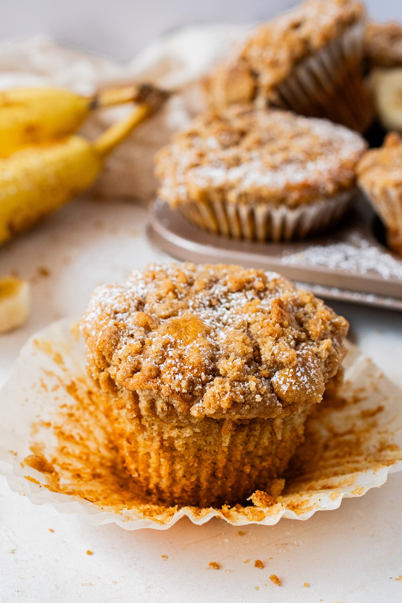 Single banana crumb muffin topped with buttery cinnamon crumble and powdered sugar, unwrapped to show the moist, fluffy texture inside.