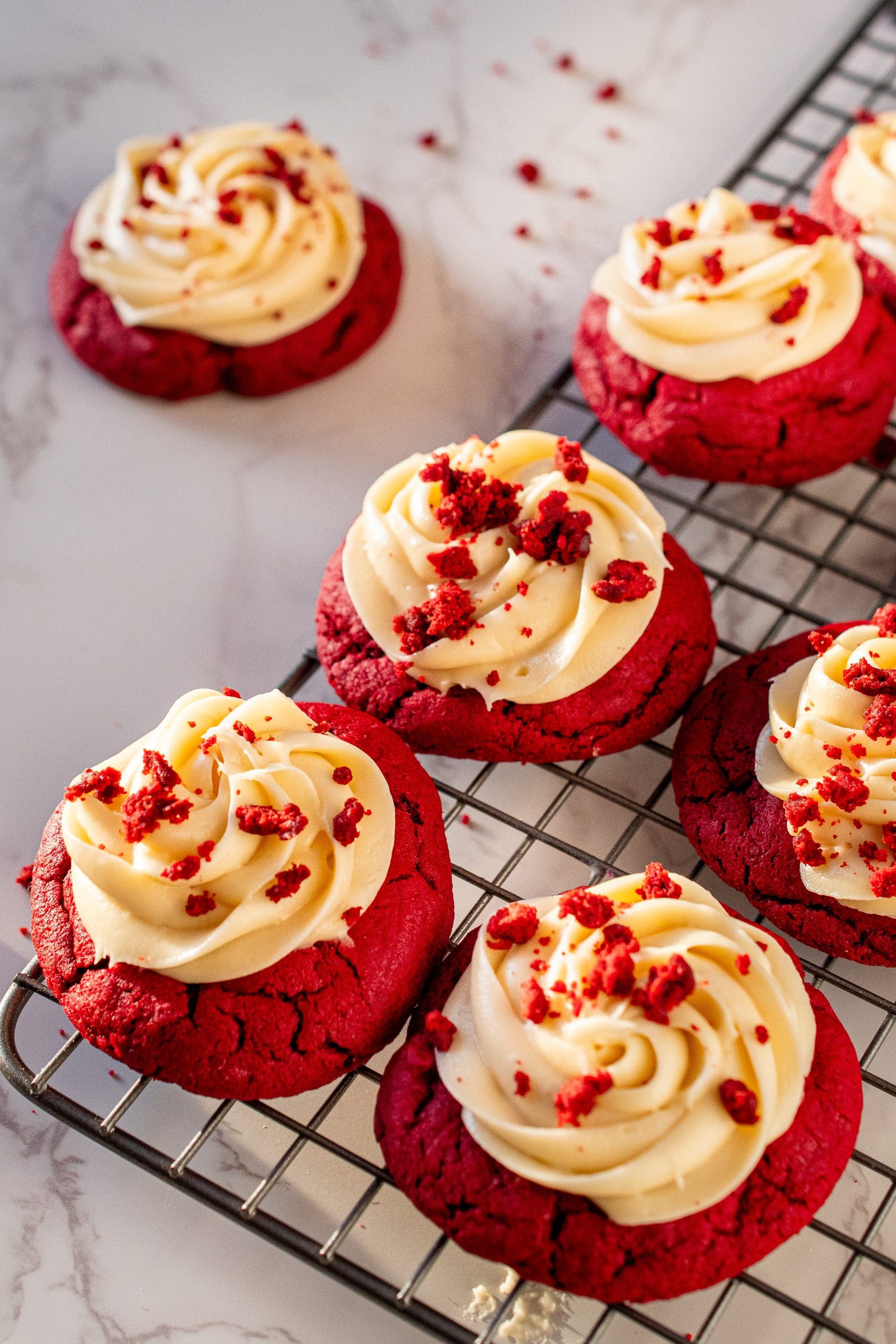Freshly baked red velvet cookies with cream cheese frosting topped with red velvet crumbs on a cooling rack.