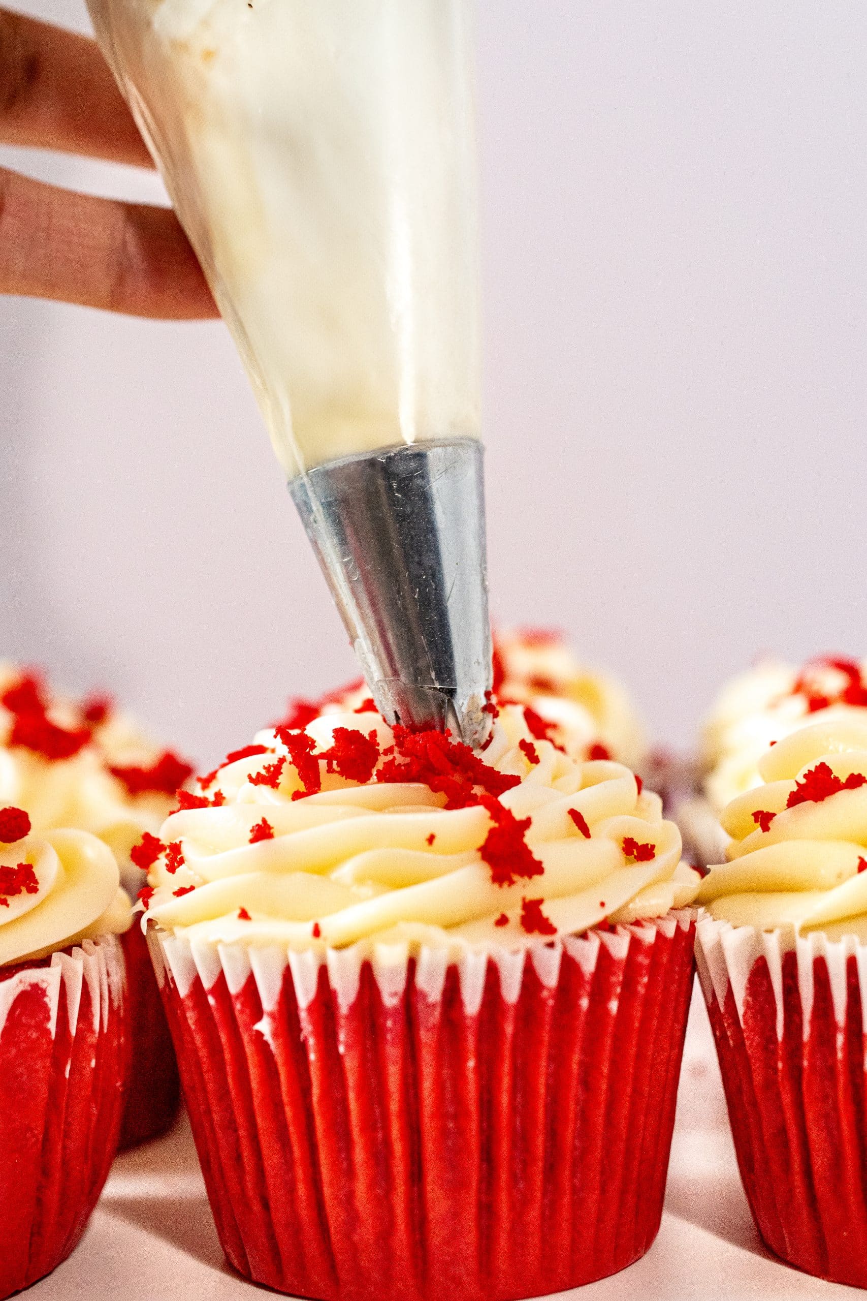 Piping cream cheese frosting onto a red velvet cupcake, with red crumbs sprinkled on top for a beautiful finish.