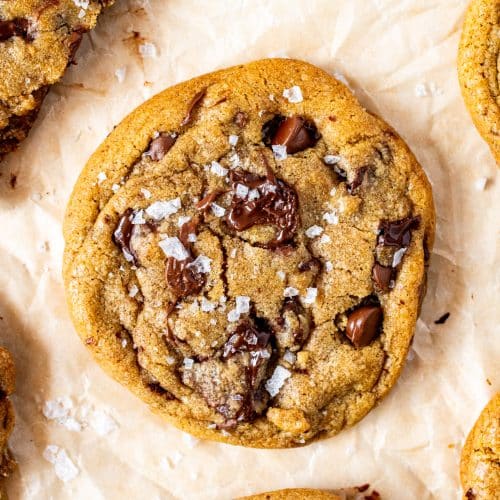 Close-up of a thick brown butter chocolate chip cookie with gooey melted chocolate chunks and a sprinkle of flaky sea salt on parchment paper.