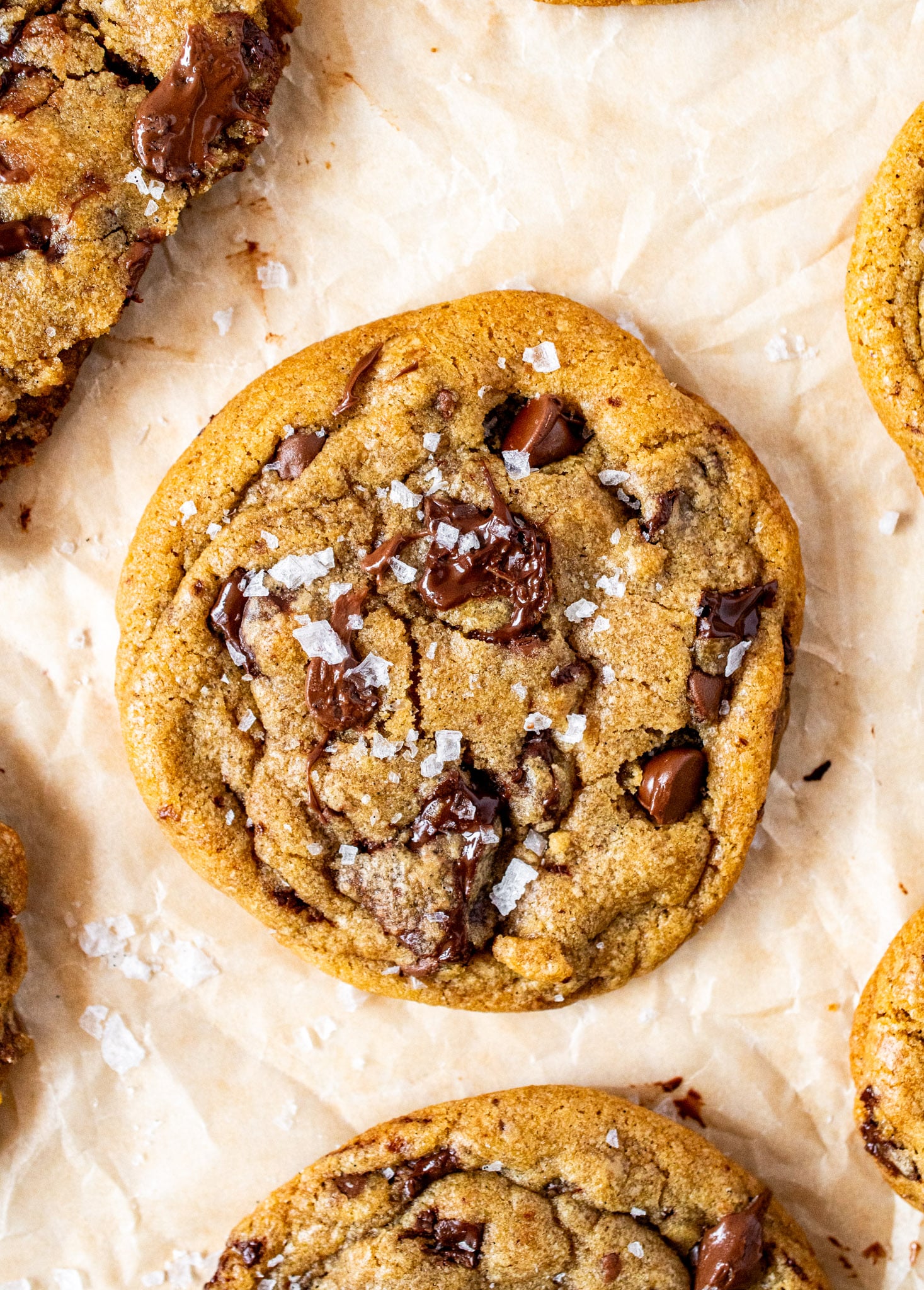 Close-up of a thick brown butter chocolate chip cookie with gooey melted chocolate chunks and a sprinkle of flaky sea salt on parchment paper.