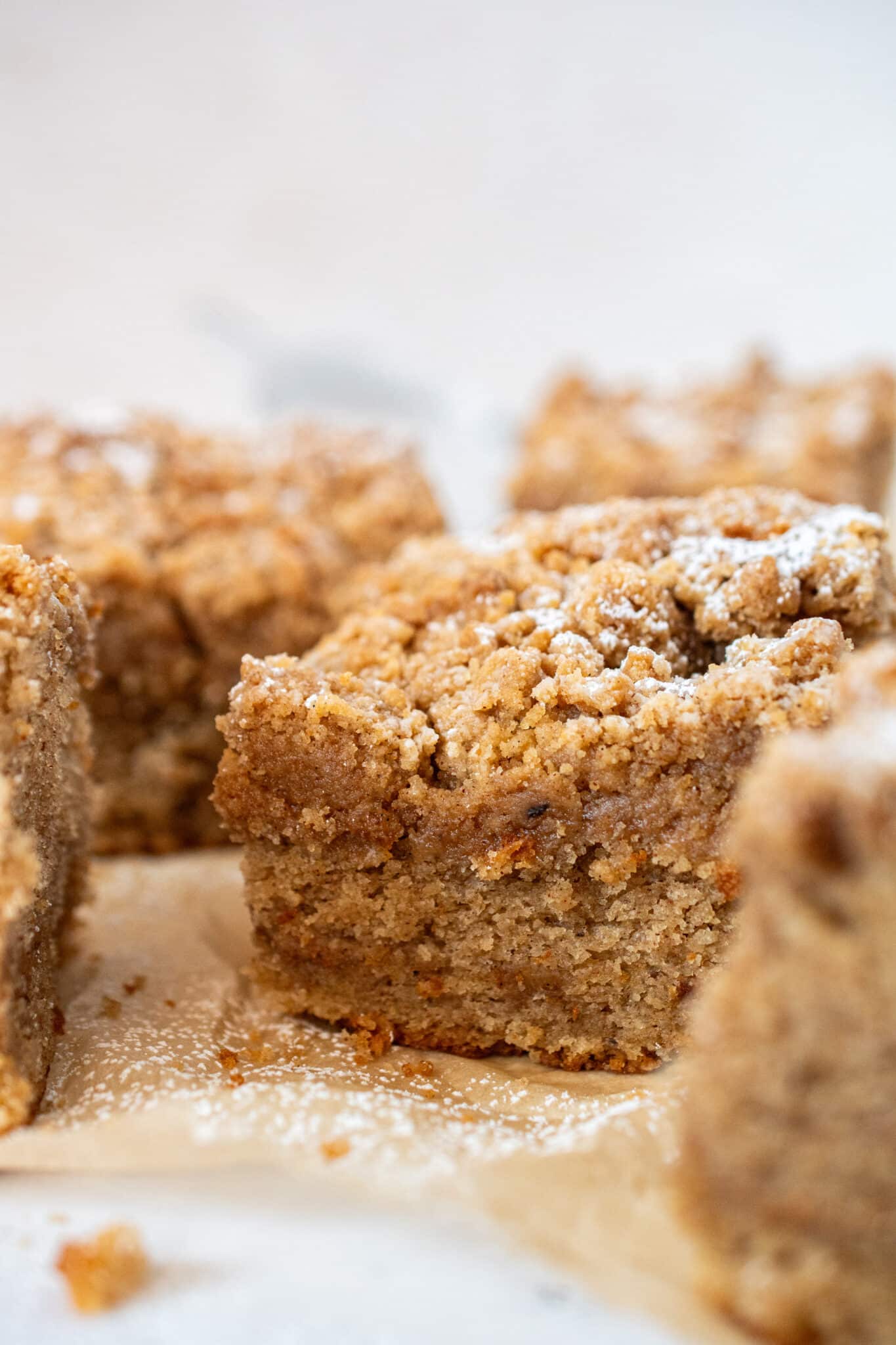Close-up of a soft and moist cinnamon crumb cake square topped with thick buttery streusel and powdered sugar on parchment paper.