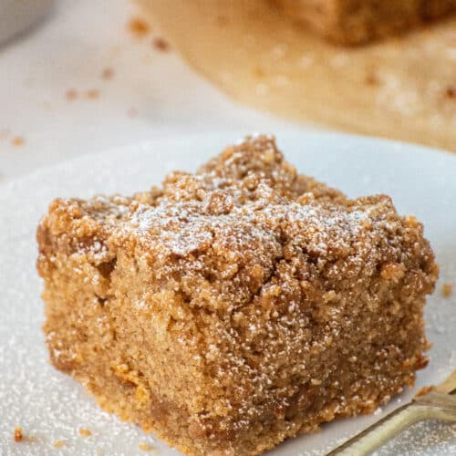 Slice of soft and moist cinnamon crumb cake topped with thick buttery streusel and powdered sugar on a white plate.