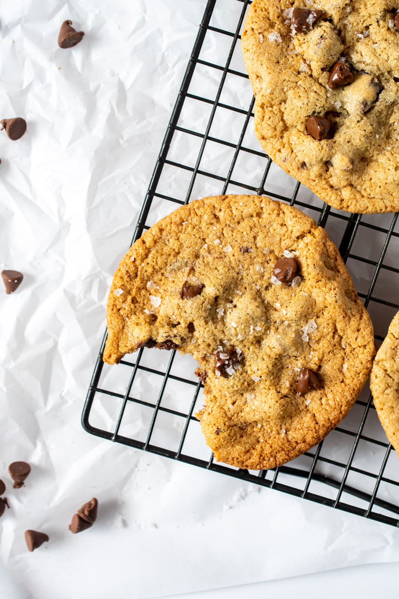 Gluten free chocolate chip cookie with crisp golden edges, chewy center, melty chocolate chips, and flaky sea salt on a black cooling rack