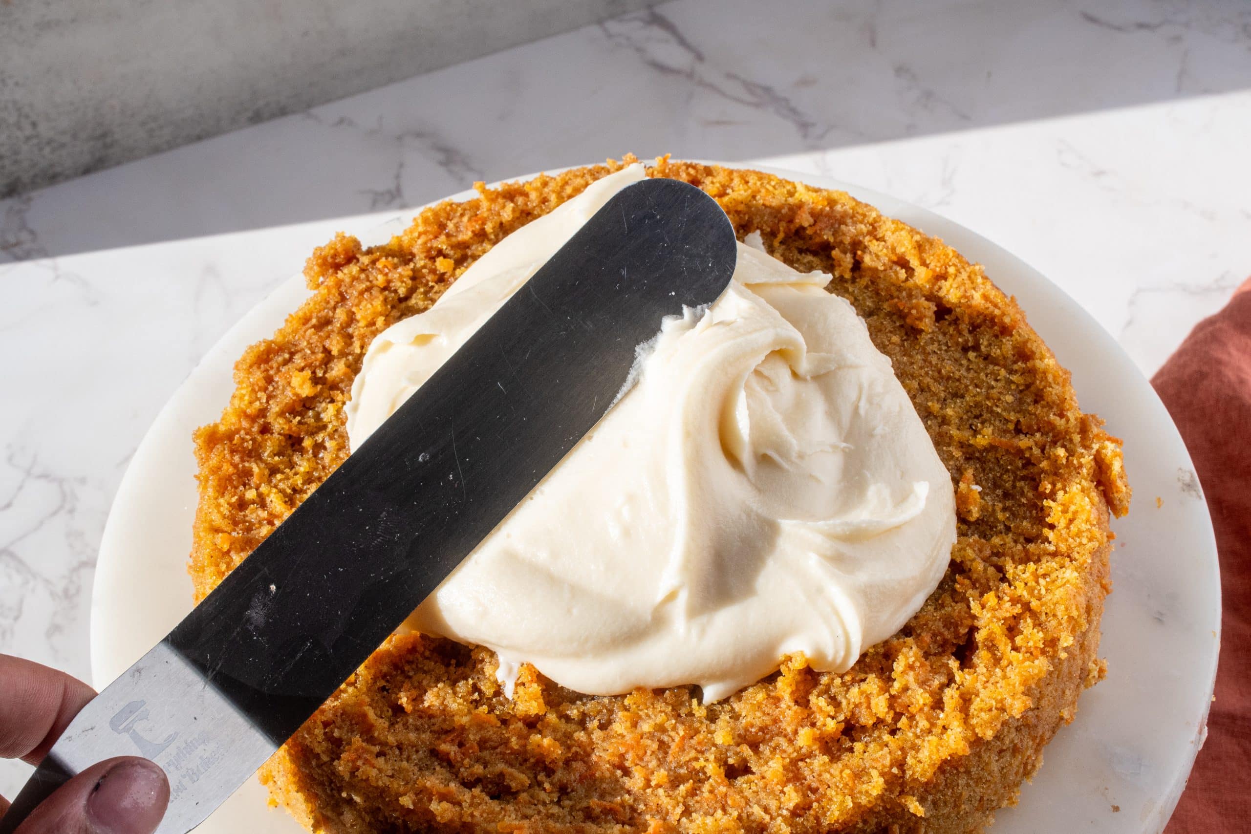 Spreading smooth cream cheese frosting on a homemade carrot cake layer using an offset spatula. The cake is golden and moist, sitting on a white plate with natural light highlighting the texture.