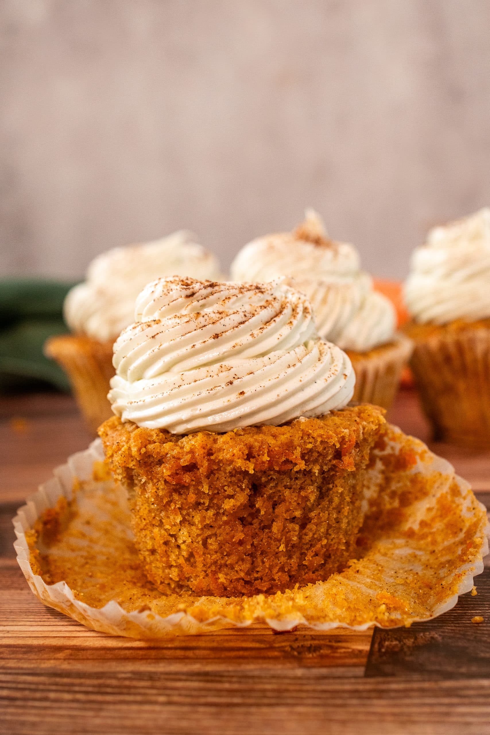 A moist carrot cake cupcake topped with creamy swirls of cream cheese frosting and a dusting of cinnamon, with more cupcakes in the background.