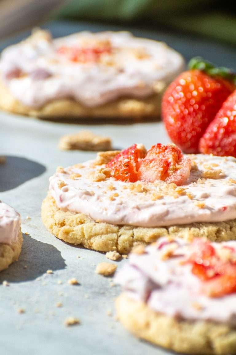 Close-up of a strawberry cheesecake cookie topped with pink cream cheese filling, sliced strawberries, and graham cracker crumbs