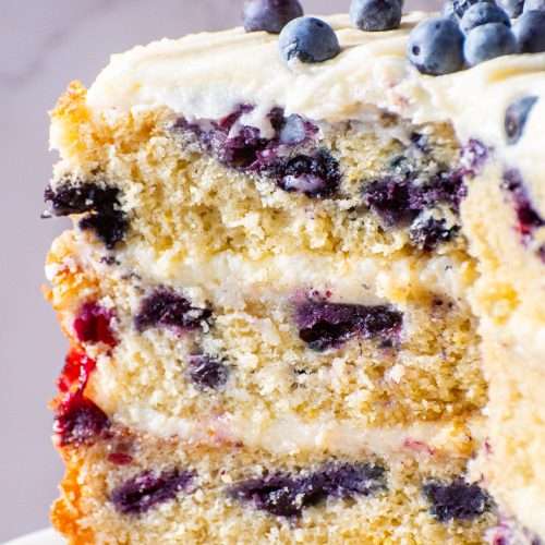 Close-up of moist lemon blueberry cake slice with layers of cream cheese frosting and juicy baked blueberries