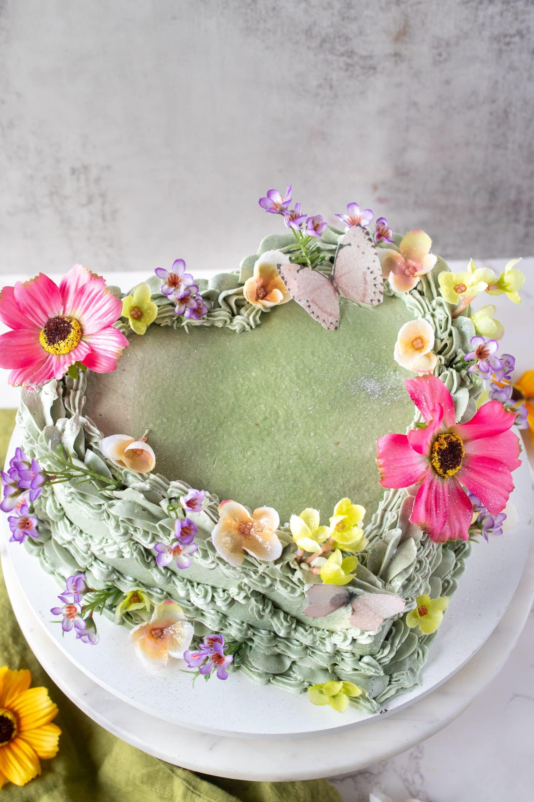 Top view of a vintage-inspired heart cake decorated with green buttercream, piped ruffles, fresh edible flowers, and delicate sugar butterflies.