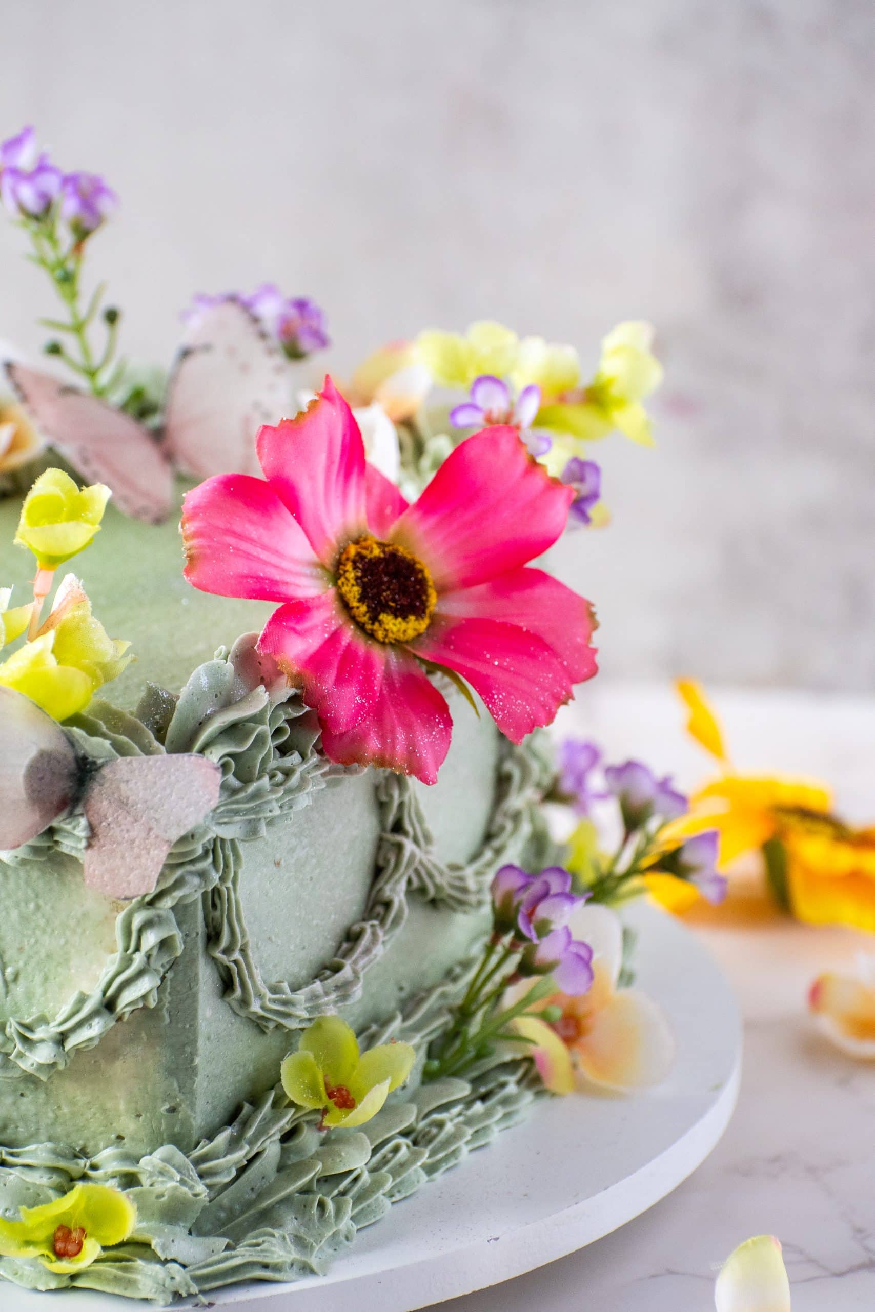 Close-up of a vintage heart cake decorated with green buttercream piping, fresh pink and yellow edible flowers, and delicate sugar butterflies.