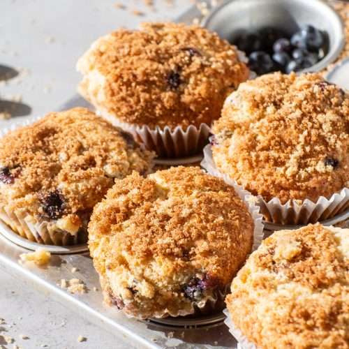 Freshly baked bakery-style blueberry muffins in a muffin tin, topped with golden brown streusel crumble.