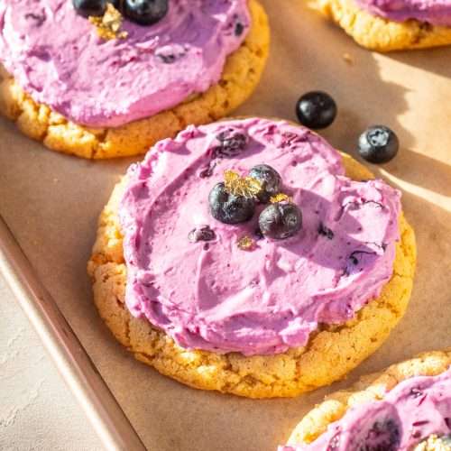 Close-up of a blueberry cheesecake cookie topped with whipped blueberry filling, fresh blueberries, and edible gold flakes on parchment paper