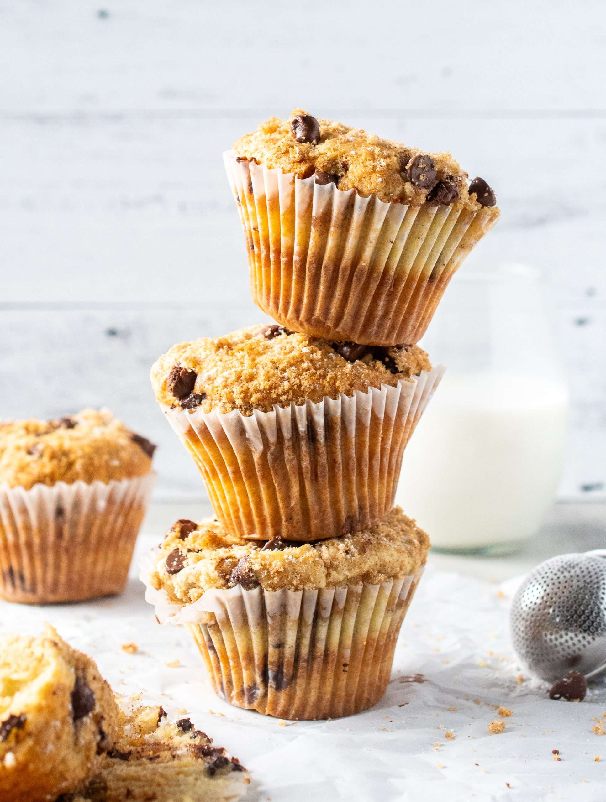 Three bakery-style chocolate chip muffins stacked on top of each other with a glass of milk in the background.