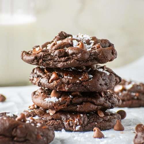 Stack of thick and chewy double chocolate chip cookies topped with flaky sea salt on parchment paper