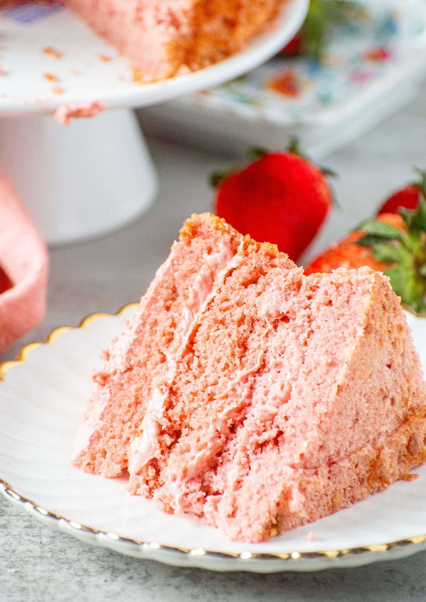 Slice of strawberry cake with creamy strawberry cream cheese frosting on a white plate with fresh strawberries in the background.