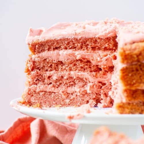 Strawberry cake with strawberry cream cheese frosting sliced on a cake stand, showing four fluffy pink layers.