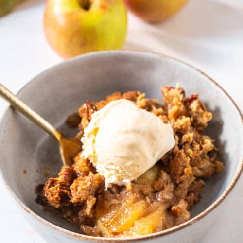 A warm bowl of apple crisp with oats and vanilla bean ice cream melting on top, served with a gold spoon.