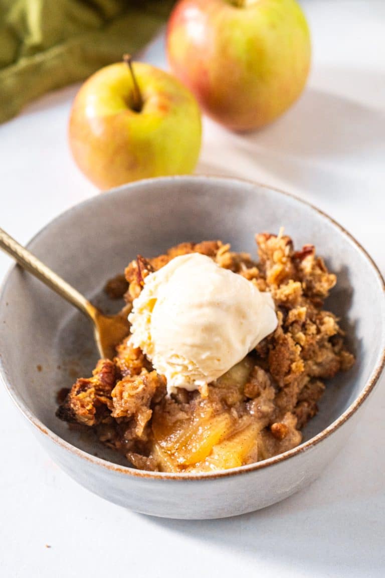 A warm bowl of apple crisp with oats and vanilla bean ice cream melting on top, served with a gold spoon.