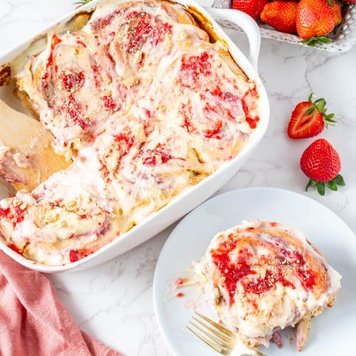 Baked strawberry cheesecake cinnamon rolls in a white dish with one roll served on a plate, topped with glaze and strawberry jam