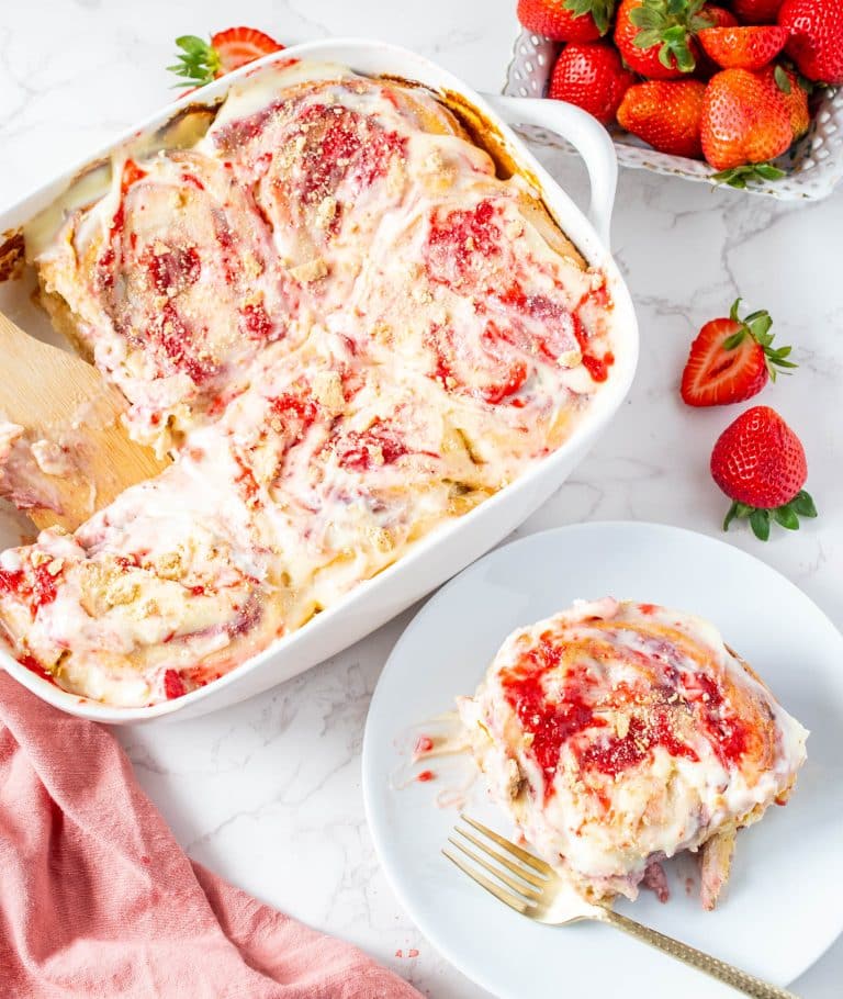 Baked strawberry cheesecake cinnamon rolls in a white dish with one roll served on a plate, topped with glaze and strawberry jam