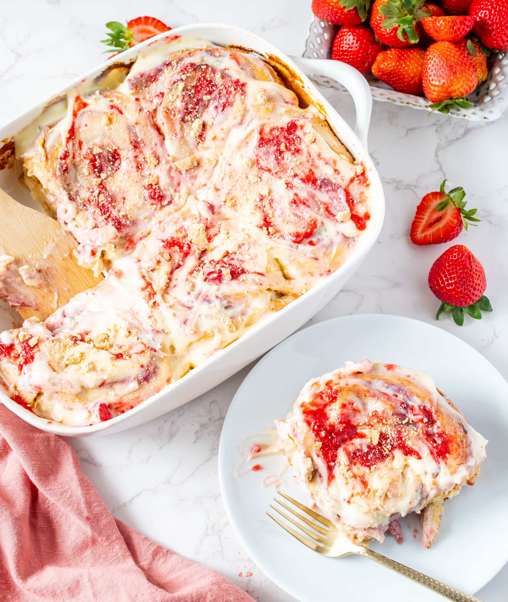 Baked strawberry cheesecake cinnamon rolls in a white dish with one roll served on a plate, topped with glaze and strawberry jam