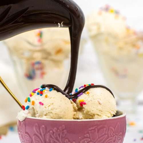 Pouring hot fudge sauce over scoops of no churn vanilla ice cream in a pink bowl topped with rainbow sprinkles.