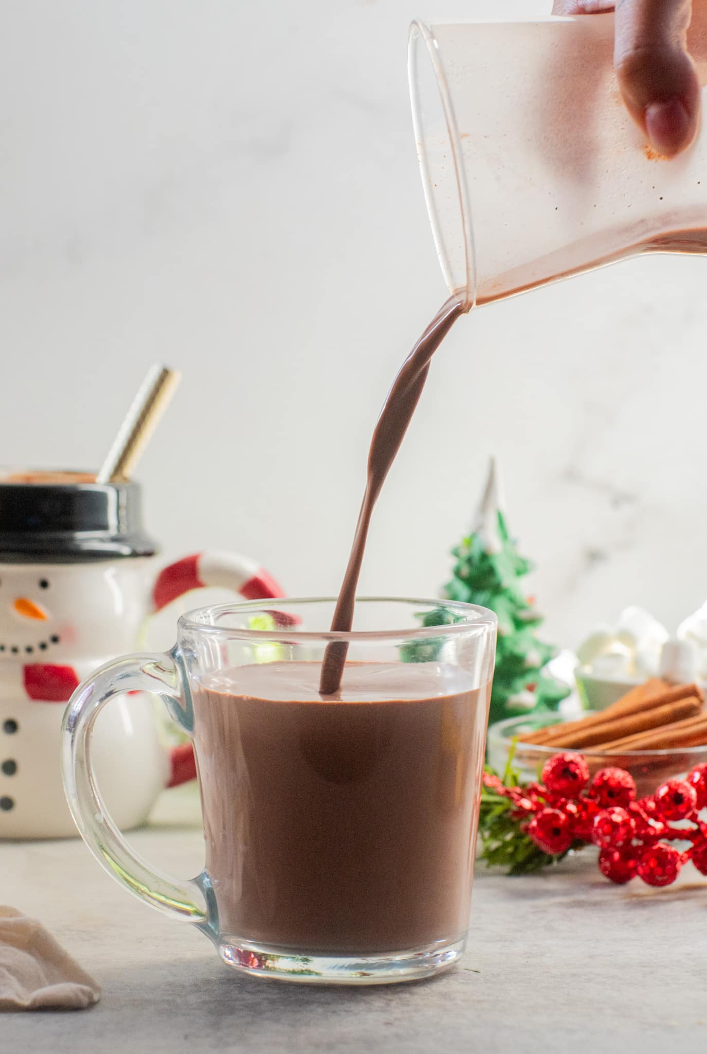 Pouring creamy hot chocolate into a glass mug surrounded by Christmas decorations, marshmallows, and a festive snowman mug in the background.