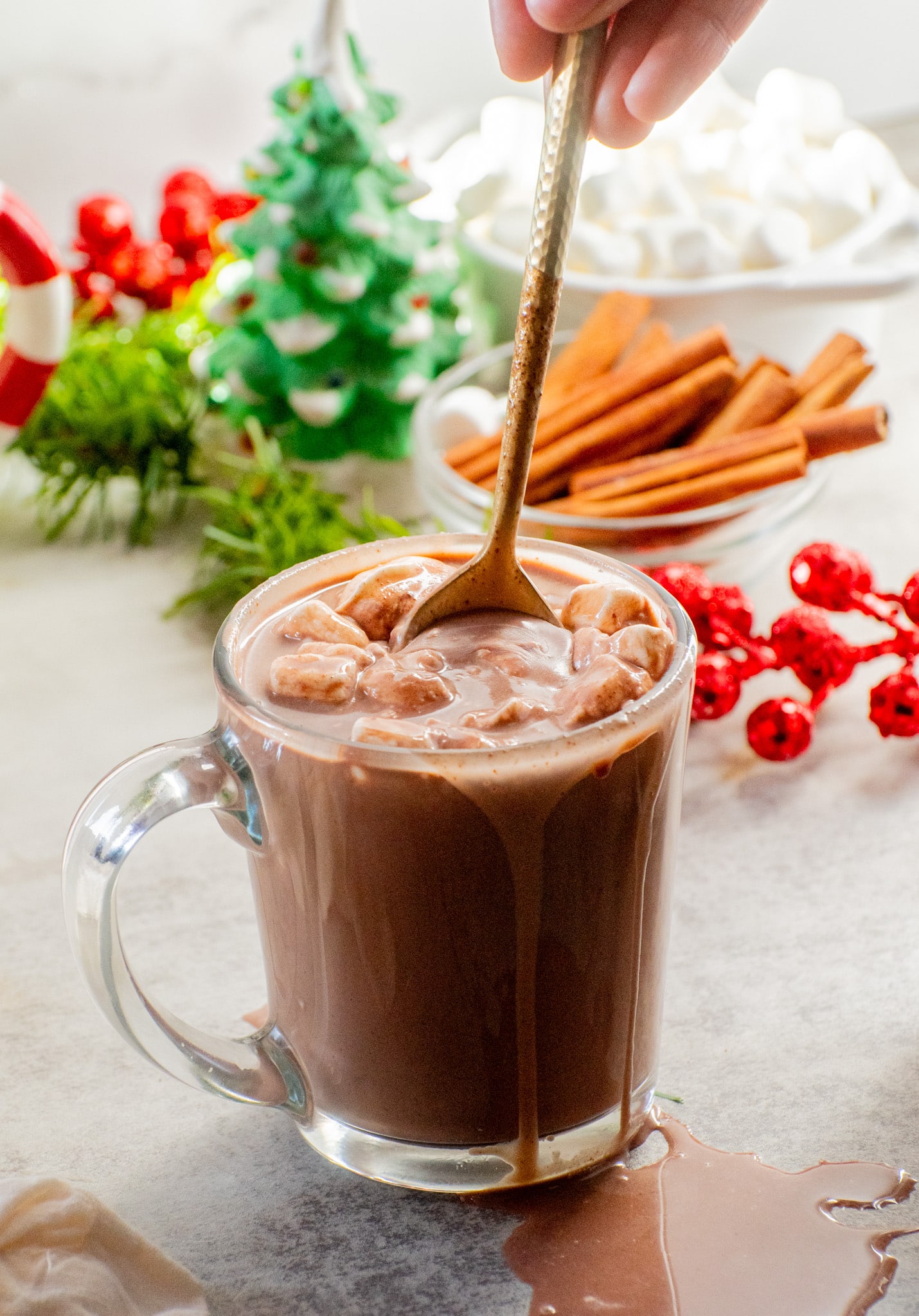 A mug of creamy hot chocolate topped with melted marshmallows and a gold spoon, surrounded by cinnamon sticks and festive Christmas décor.