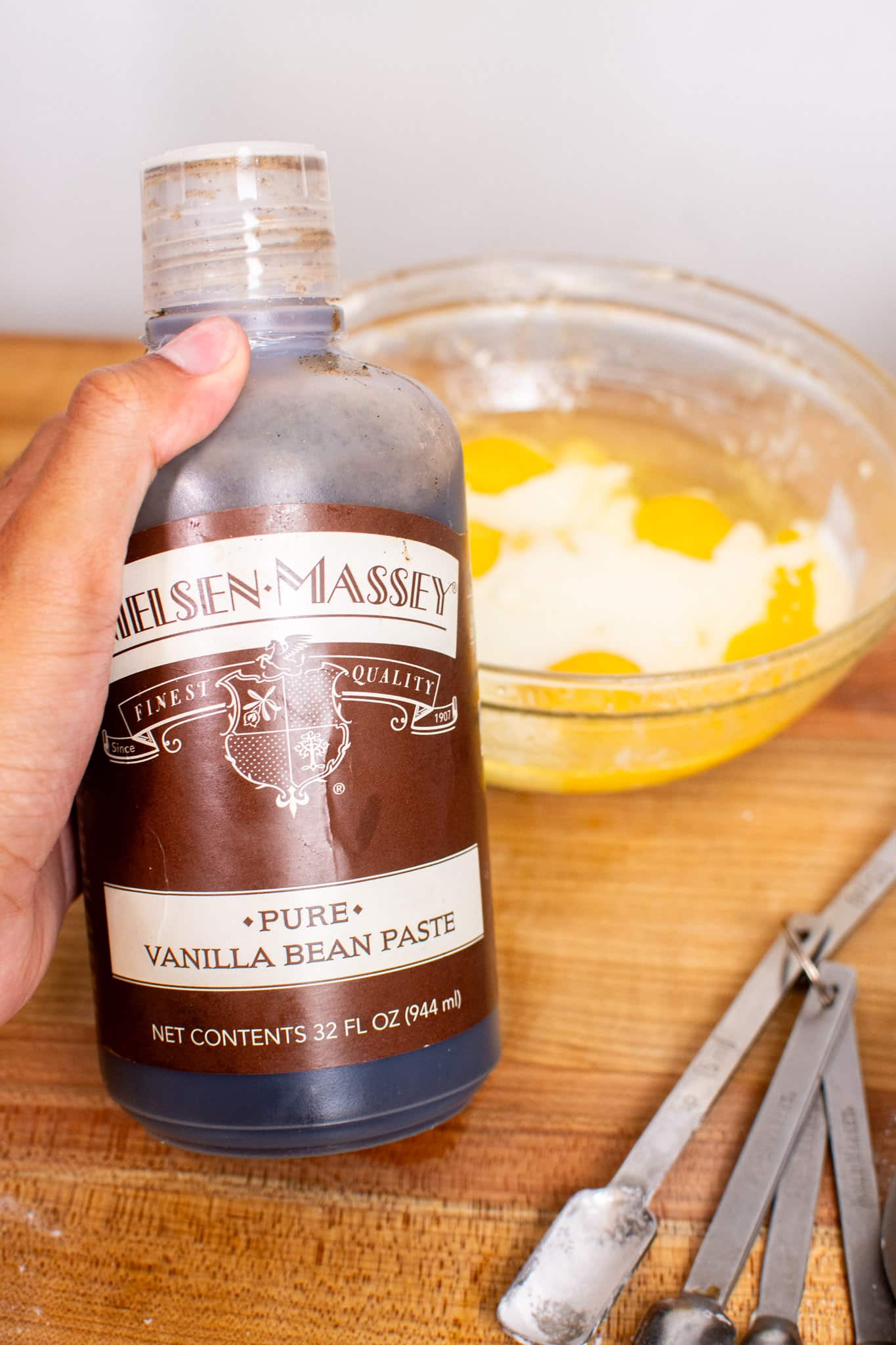 Hand holding Nielsen-Massey pure vanilla bean paste bottle with eggs in a mixing bowl in the background