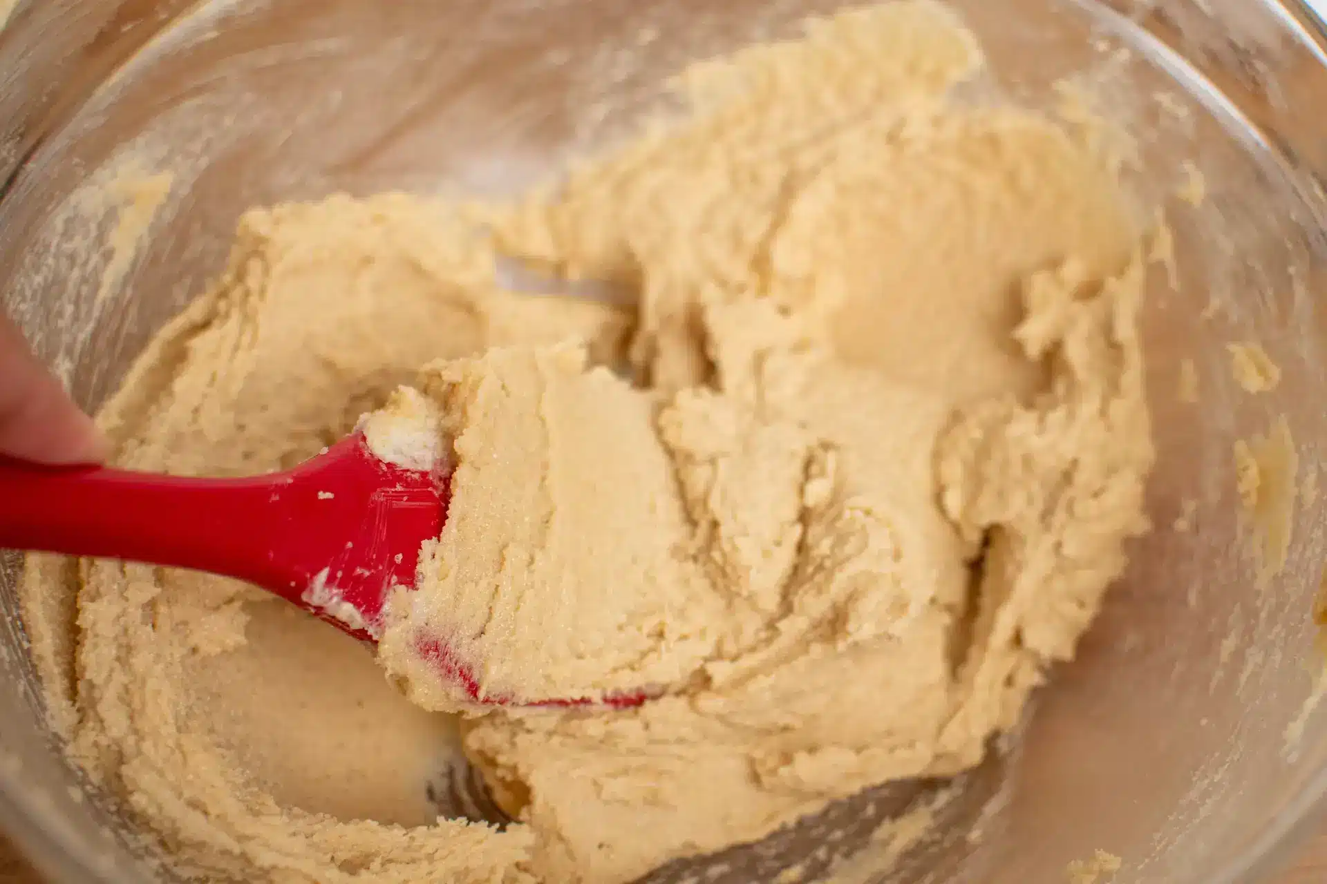 Cookie dough being mixed with a red spatula in a glass bowl.