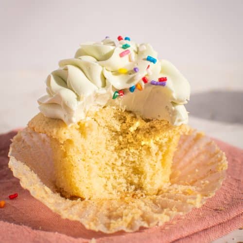 Close-up of a vanilla cupcake with a bite taken out, topped with fluffy vanilla frosting and colorful sprinkles, displayed on a pink napkin.
