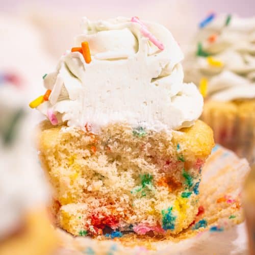 Close-up of a sliced Funfetti cupcake with colorful sprinkles and a swirl of vanilla buttercream frosting on top.