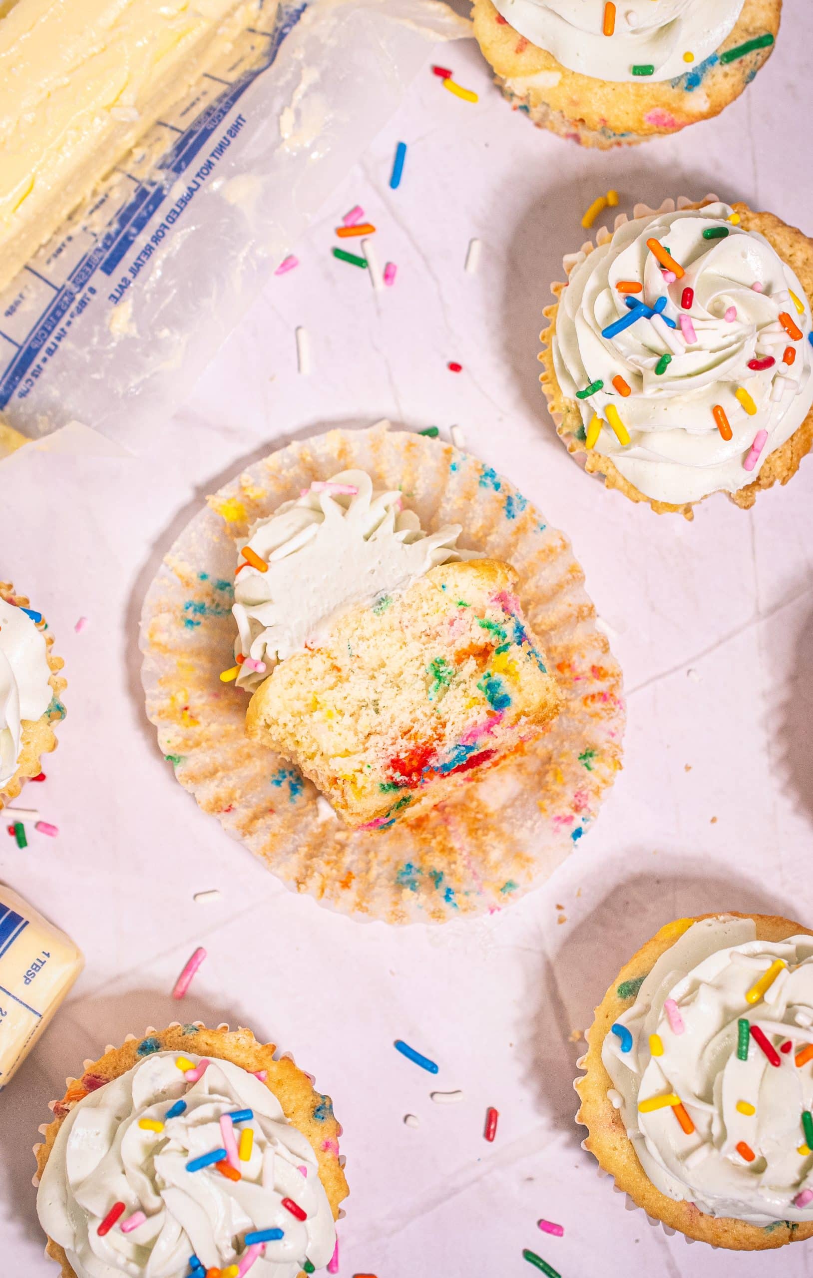 Top-down view of a sliced Funfetti cupcake with vanilla buttercream frosting and colorful sprinkles, with cupcake wrapper opened.