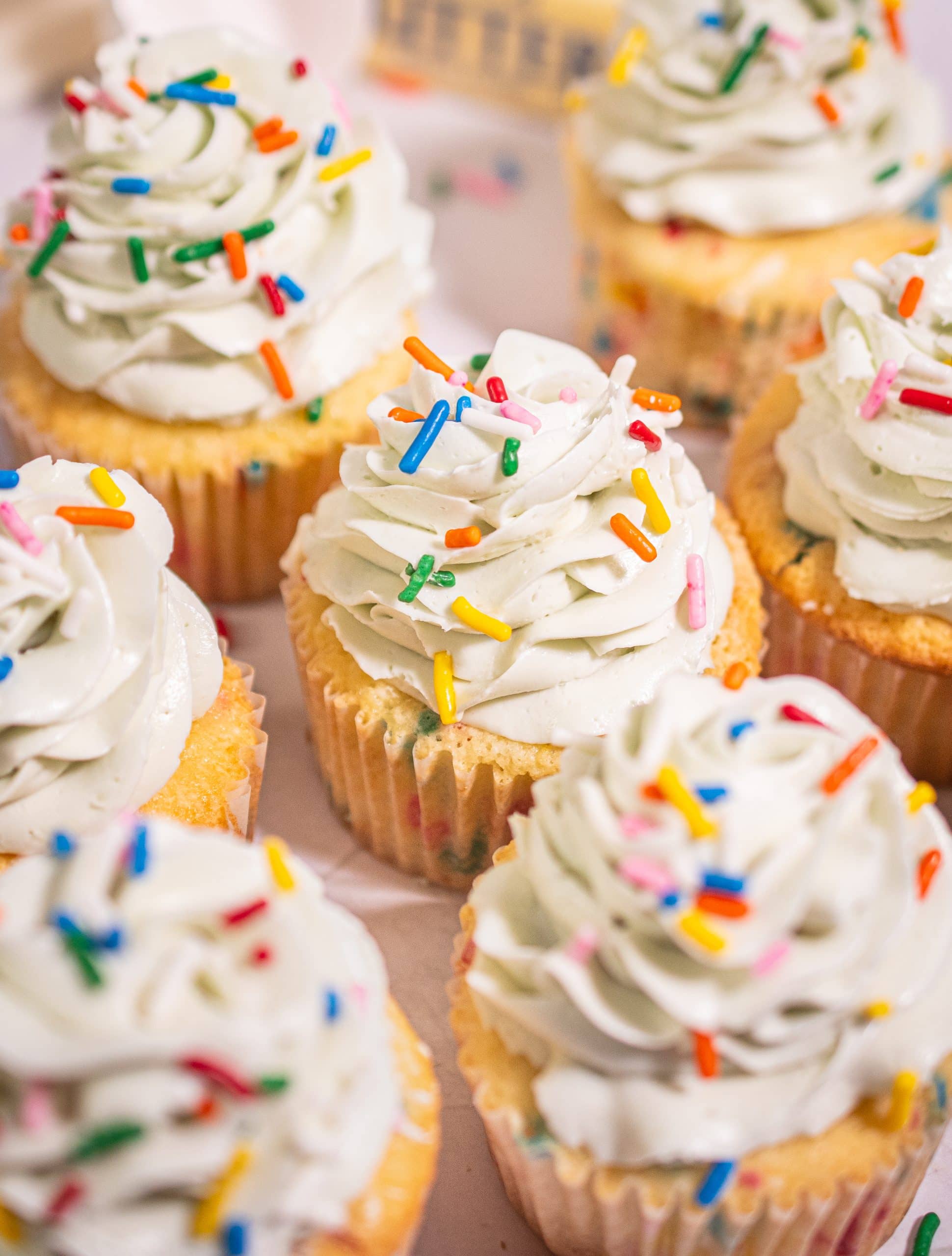 Top-down view of Funfetti cupcakes with vanilla buttercream frosting and colorful sprinkles on a pink background, with one cupcake partially unwrapped.