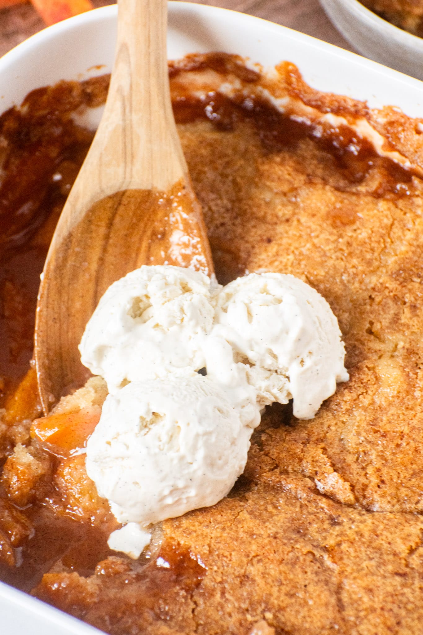 Baking dish of homemade peach cobbler topped with three scoops of vanilla ice cream on a golden brown crust
