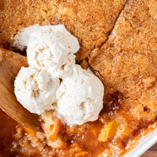 Spoonful of homemade peach cobbler in baking dish with scoops of vanilla ice cream on top