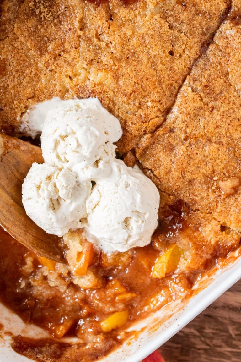 Spoonful of homemade peach cobbler in baking dish with scoops of vanilla ice cream on top