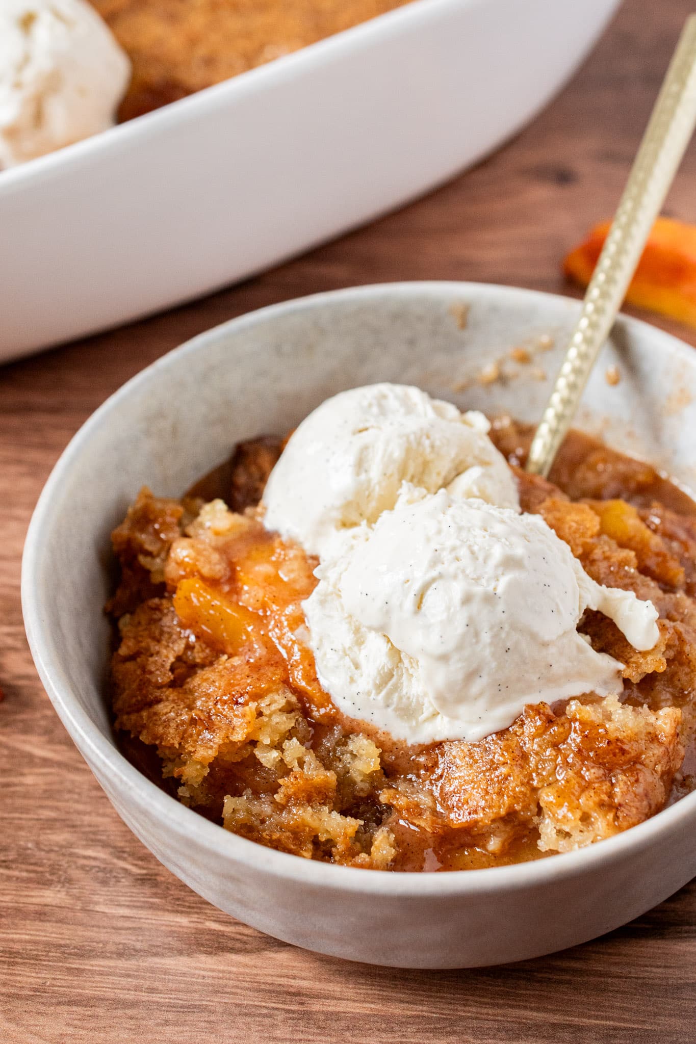 Bowl of homemade peach cobbler topped with two scoops of melting vanilla ice cream and a golden spoon
