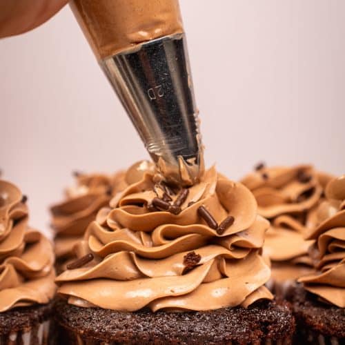 Close-up of piping chocolate buttercream frosting onto a chocolate cupcake topped with sprinkles.
